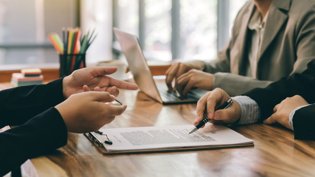 Three people looking over paperwork, one holding a pen and one typing on a laptop sat at a wooden table