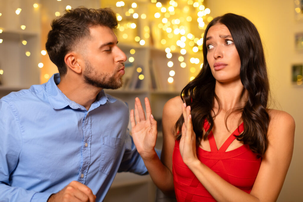 Man leans in puckering for kiss, making a playful expression, while the woman appears unimpressed, rejecting his advance with a hand gesture