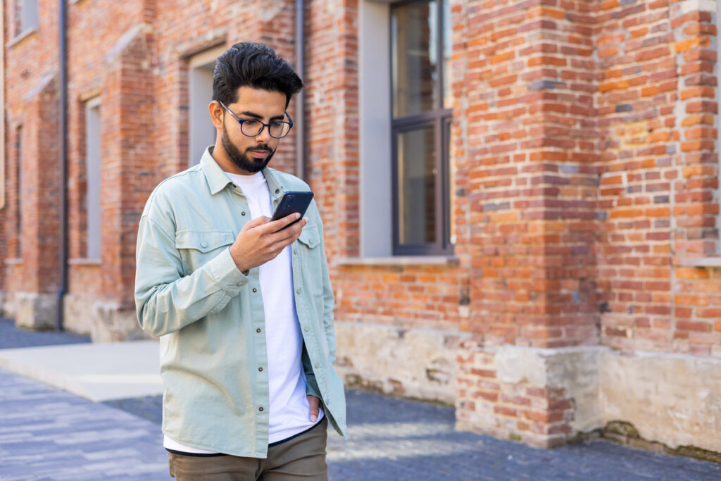 A man walks down the street with one hand in his pocket and then other holding his phone up to look at it