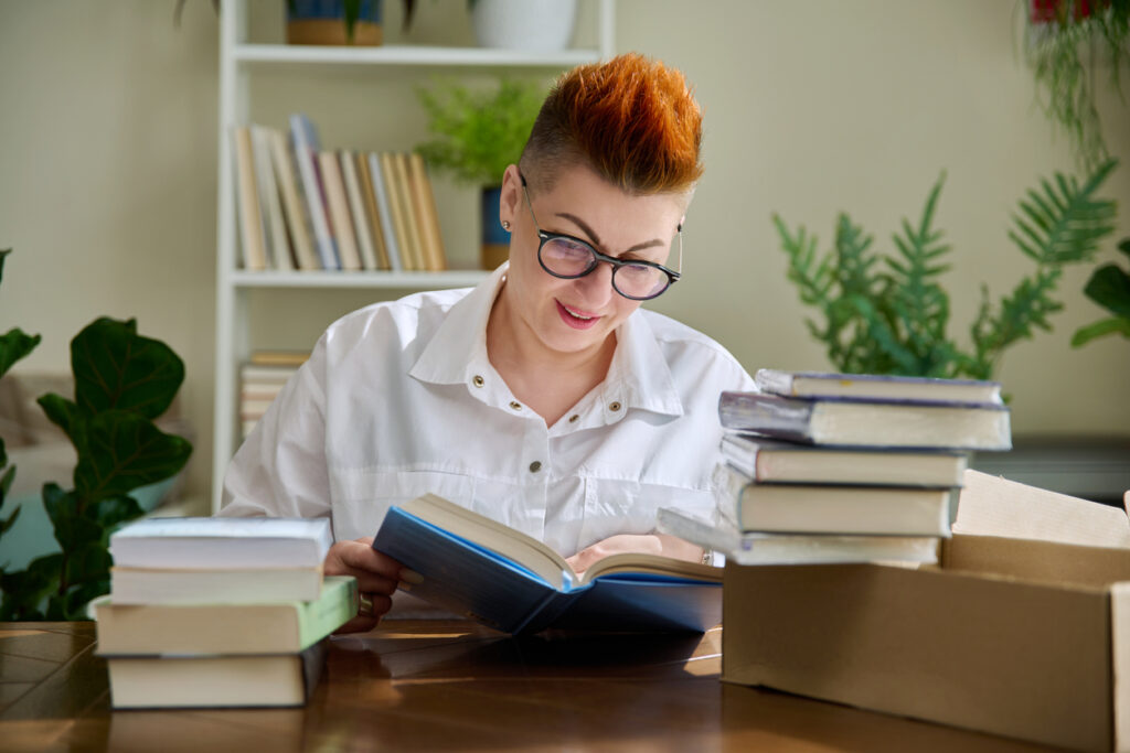 Woman with new books unpacked from cardboard box from an online store. Plants and a book shelf in the background