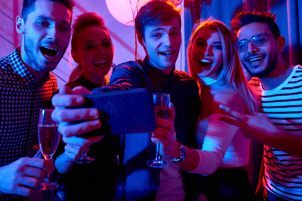 Group of smiling young people taking selfie looking at smartphone camera while enjoying a celebration